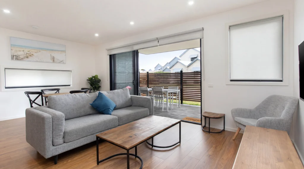 Spacious ground floor apartment lounge room in Gippsland featuring comfortable sofas, flat-screen TV, and large windows overlooking native gardens